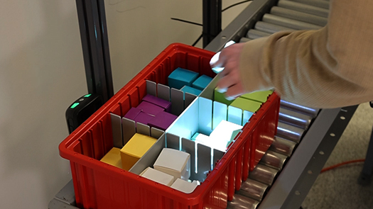 A hand is reaching into a divided tote on a conveyor, being guided by a projection picking system.